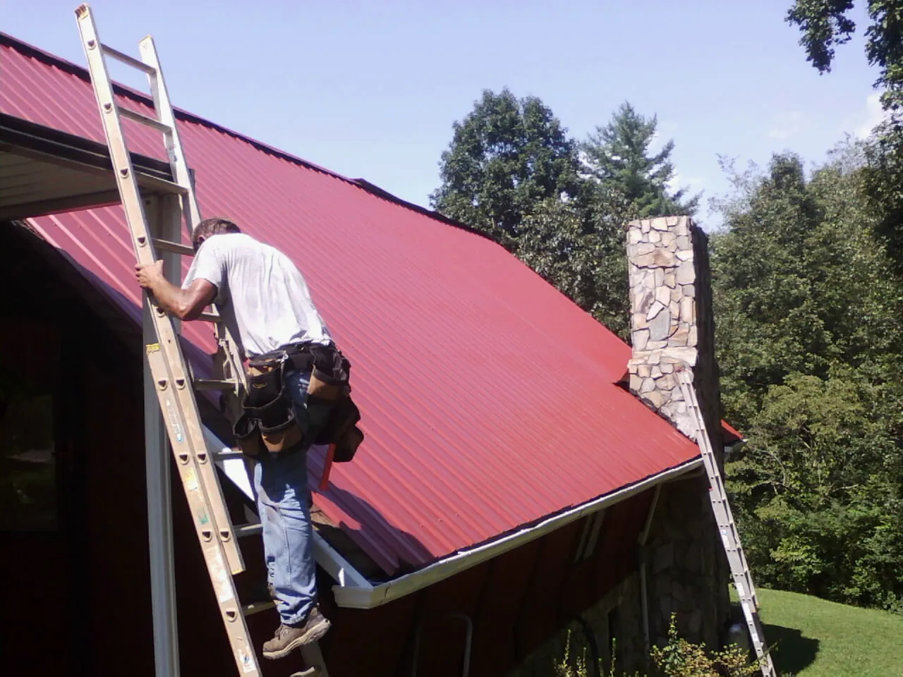 Roofer on a ladder beside a red metal roof and stone chimney — Soffit & Fascia Repair by Roofing Brothers in Holmdel